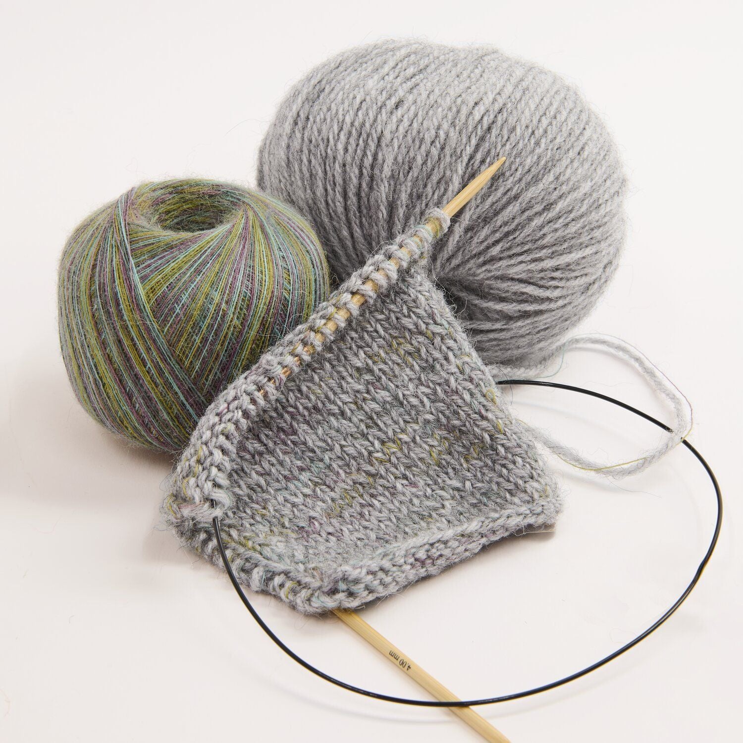 A close-up studio shot of knitting materials on a white background: a gray knitted swatch on a wooden circular needle, one ball of light gray yarn, and one ball of variegated yarn with green, blue, and purple tones.