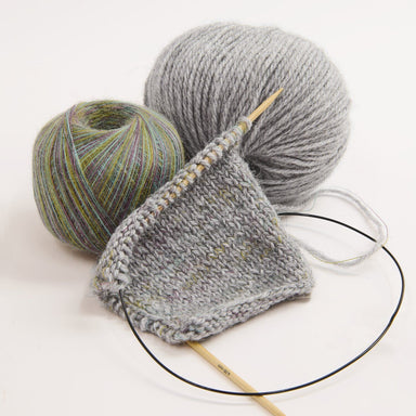 A close-up studio shot of knitting materials on a white background: a gray knitted swatch on a wooden circular needle, one ball of light gray yarn, and one ball of variegated yarn with green, blue, and purple tones.