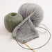 A close-up studio shot of knitting materials on a white background: a gray knitted swatch on a wooden circular needle, one ball of light gray yarn, and one ball of variegated yarn with green, blue, and purple tones.