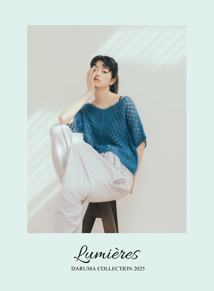 Woman wearing a blue knit sweater and white trousers against a neutral background with 'Lumières Daruma Collection 2025' text.