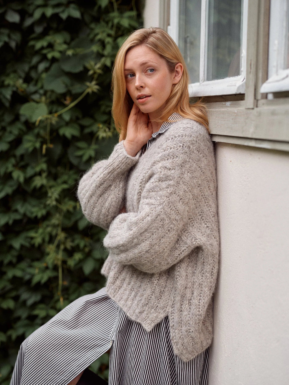 Woman wearing a chunky knit sweater leaning against a building with greenery in the background