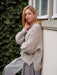 Woman wearing a chunky knit sweater leaning against a building with greenery in the background
