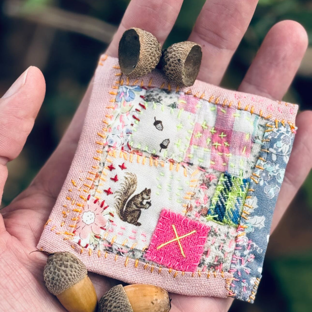 Hand holding a small patchwork fabric square with acorns on a blurred natural background