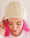 A close-up of a person wearing a cream cable-knit beanie hat, with bright pink hair and a white top against a plain light background.
