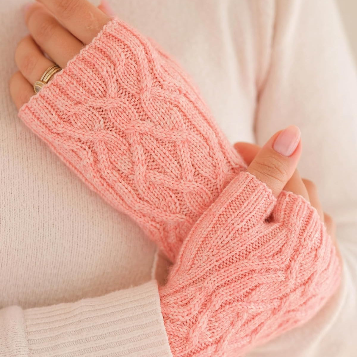 A close-up of a person wearing light pink cable-knit fingerless gloves over a cream-colored knit sweater.