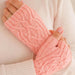 A close-up of a person wearing light pink cable-knit fingerless gloves over a cream-colored knit sweater.