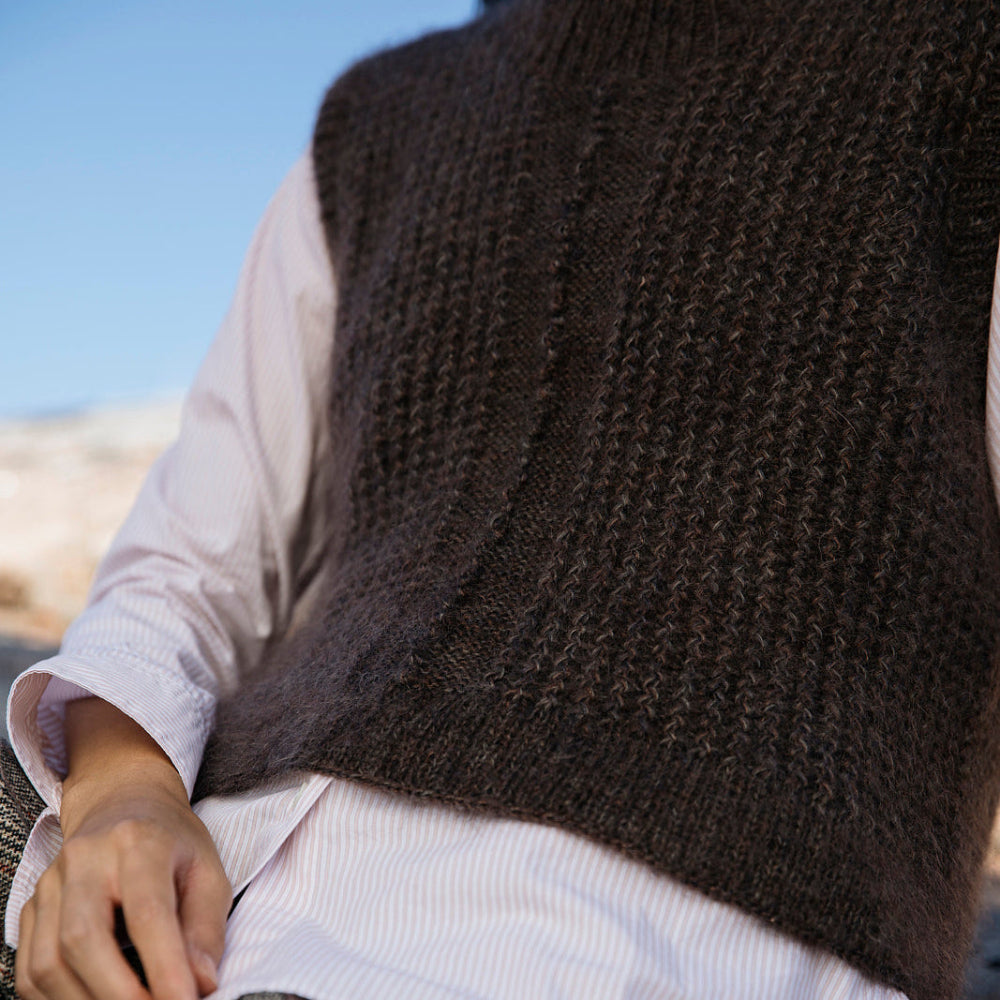 Person wearing a brown knitted vest over a white shirt with plaid pants.