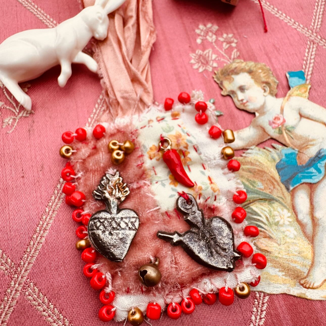 Decorative heart with red beads, silver charms, and a small angel figure on a pink fabric background.