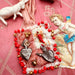 Decorative heart with red beads, silver charms, and a small angel figure on a pink fabric background.