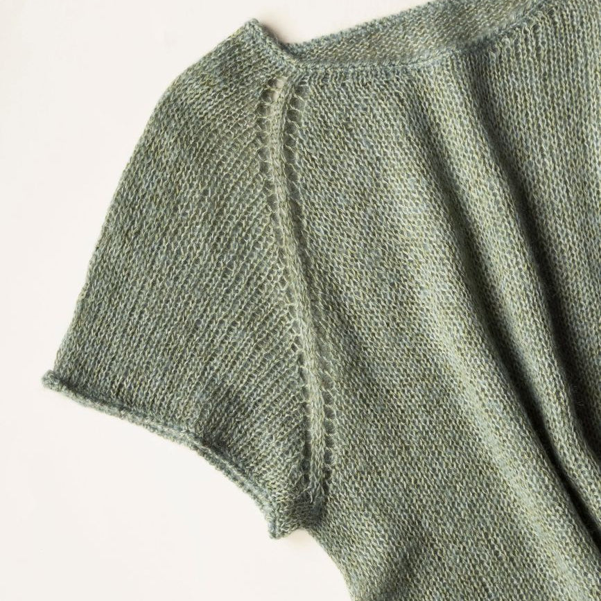 Close-up of a textured knit garment in a muted sage green color, showing the ribbed sleeve and shoulder construction against a plain off-white background.