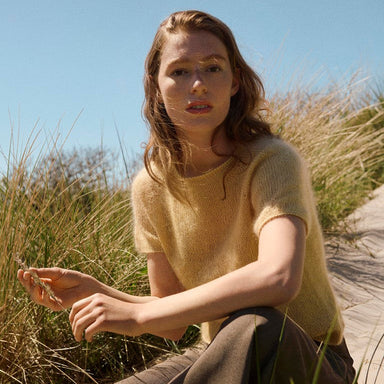 A woman sitting outdoors on a sandy path among tall grass under a clear blue sky, wearing a pale yellow short-sleeve knit top and brown wide-leg pants.