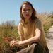 A woman sitting outdoors on a sandy path among tall grass under a clear blue sky, wearing a pale yellow short-sleeve knit top and brown wide-leg pants.