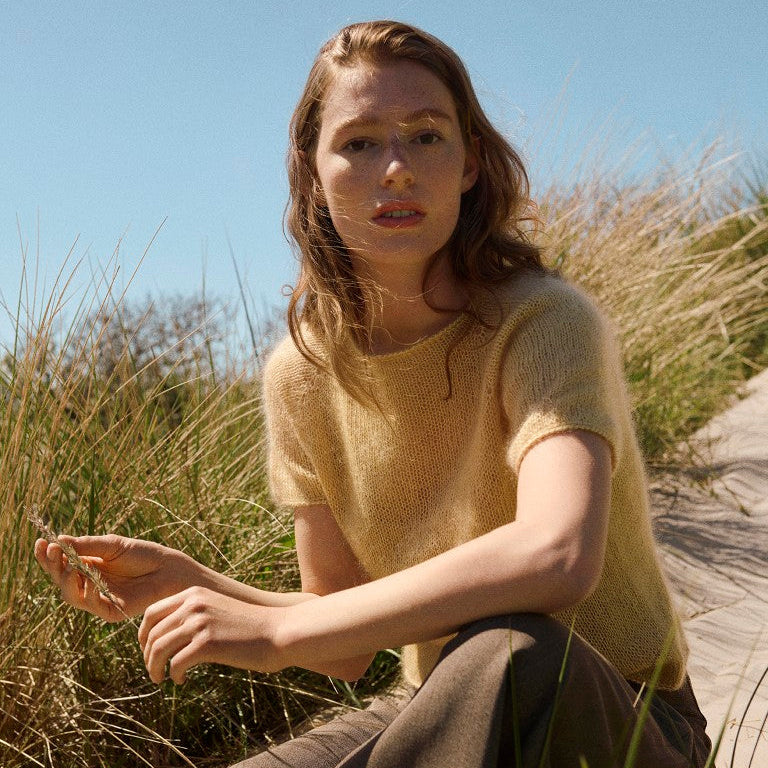 A woman sitting outdoors on a sandy path among tall grass under a clear blue sky, wearing a pale yellow short-sleeve knit top and brown wide-leg pants.