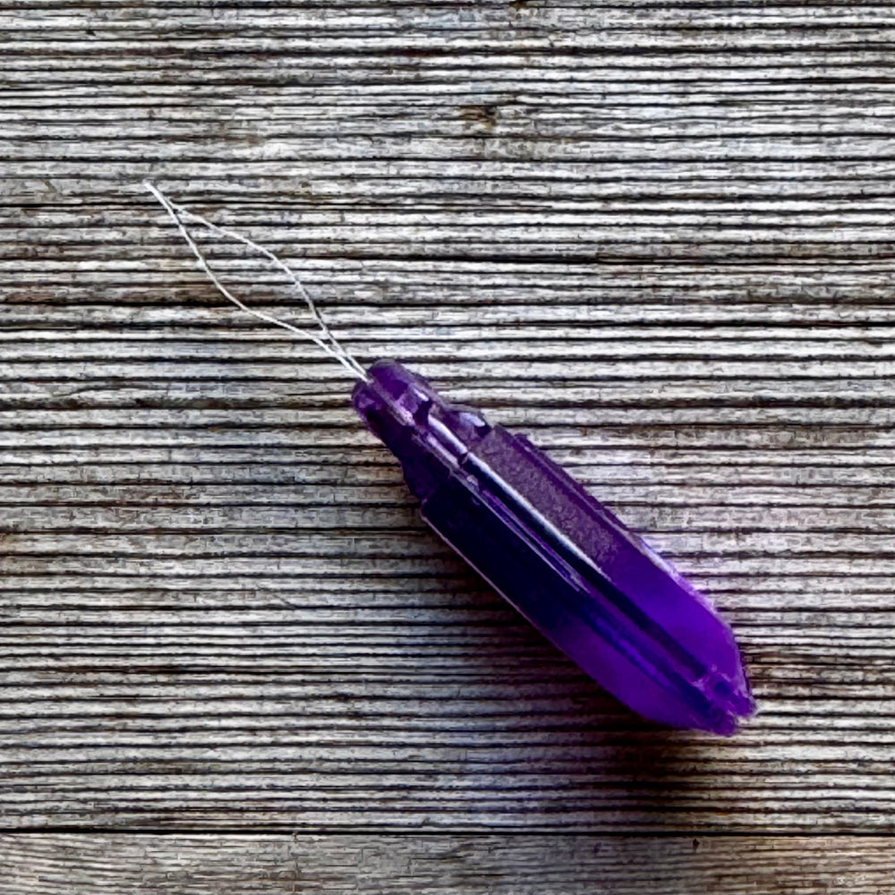 A translucent purple crystal pendant with a white thread loop, resting diagonally on a rustic, weathered gray wood surface.
