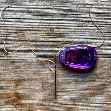 A purple translucent plastic spool of copper-colored thread lies on a weathered wooden surface, with the thread looped around a metal needle.