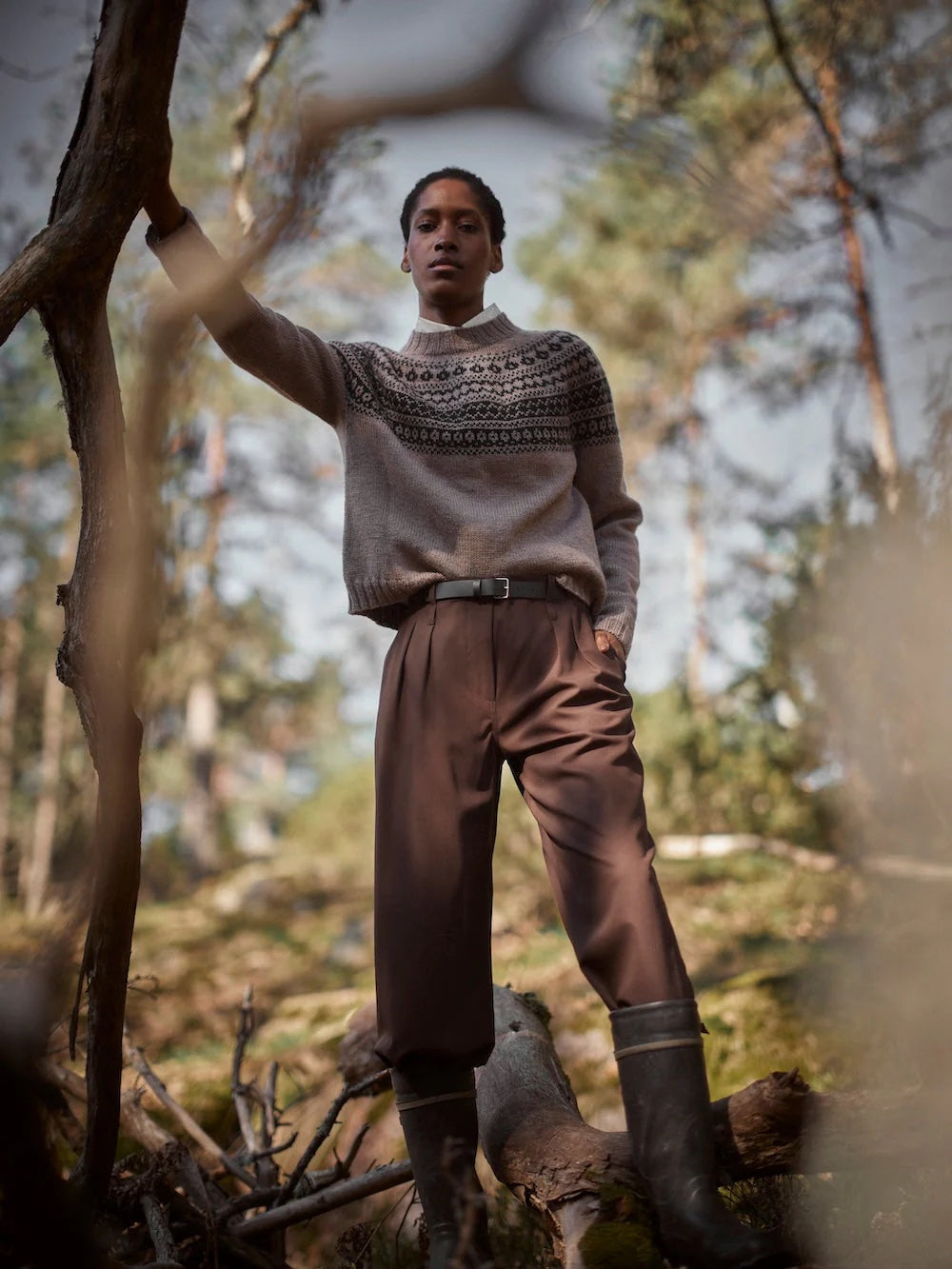 Person wearing a patterned sweater and brown pants standing in a forest.