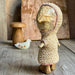 A handmade knitted doll figure with a paper face and wooden legs, standing on a rustic wooden surface with a wooden mushroom and a small white bird figurine in the background.
