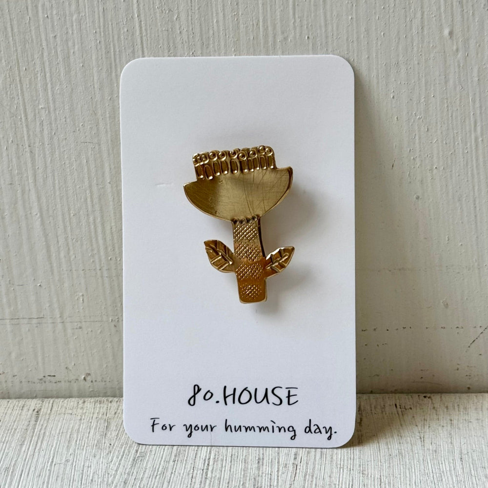 A gold metal hummingbird pin displayed on a white backing card that reads '80.HOUSE For your humming day.