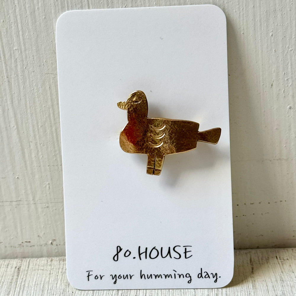 A small duck-shaped brooch in a textured gold-tone metal with a subtle red accent on the body, mounted on a white display card that reads '80.HOUSE For your humming day.