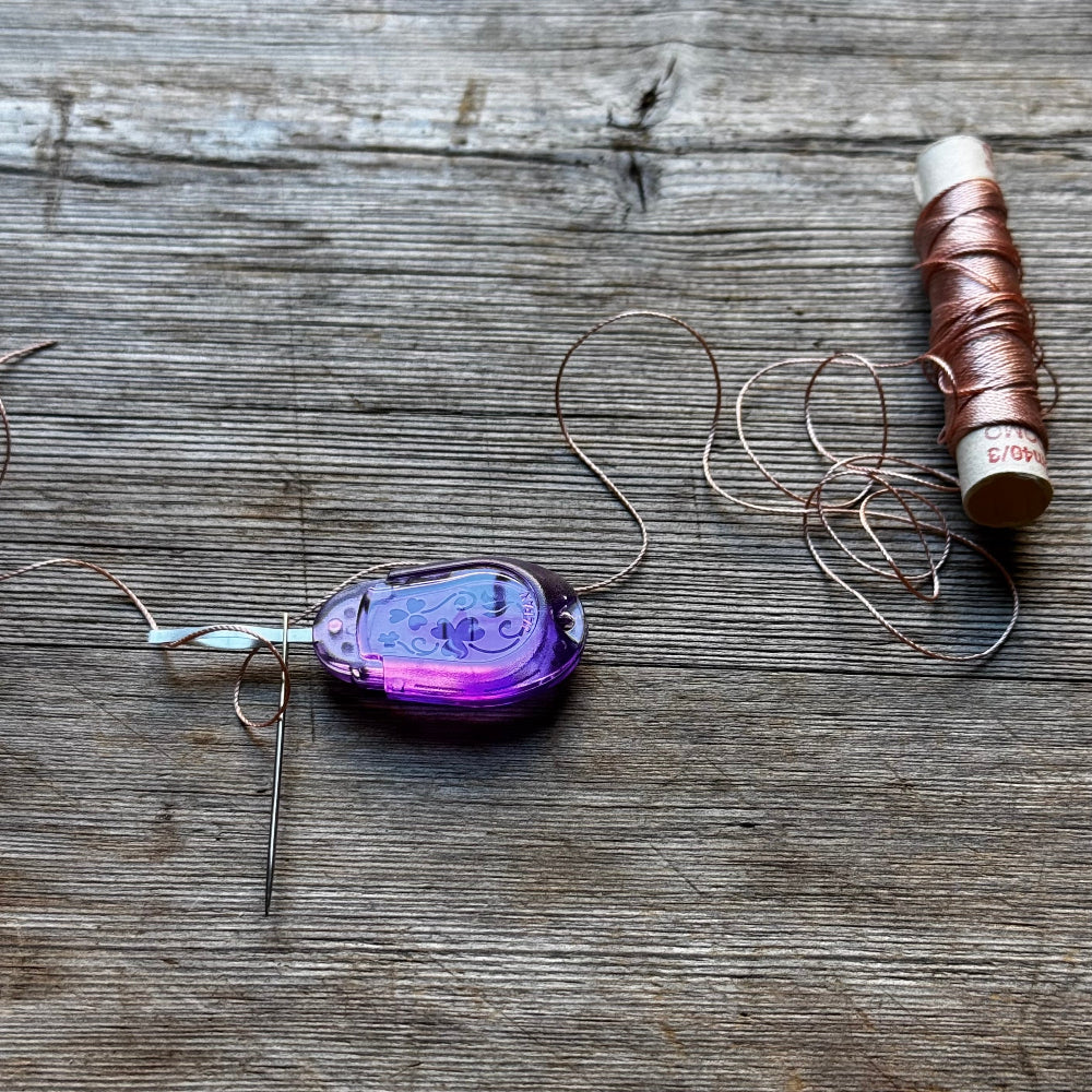 A purple translucent bead or cabochon with attached copper wire wrapped around it, placed on a rustic wooden surface next to a spool of copper thread and a needle.