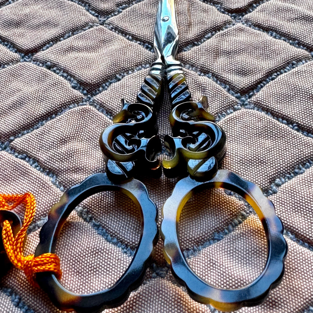 Decorative scissors with ornate dark handles and tortoiseshell-style oval blades, placed on a quilted fabric background with an orange cord loop.