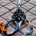 Decorative scissors with ornate dark handles and tortoiseshell-style oval blades, placed on a quilted fabric background with an orange cord loop.