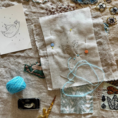 A styled flat-lay of embroidery materials on a beige linen surface, including a light blue ball of yarn, gold-handled scissors, a small tin, a printed bird sketch card, and several fabric swatches with embroidered details.