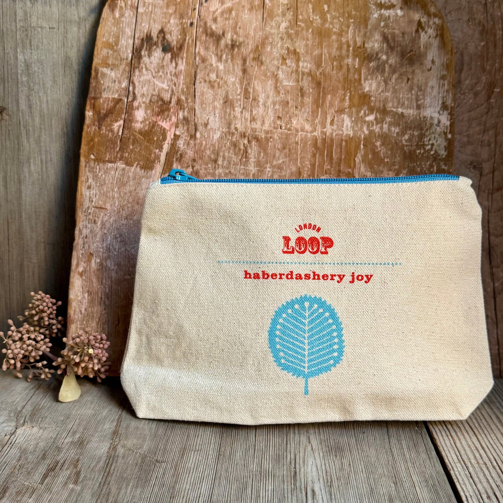 A canvas cosmetic pouch with a blue zipper, featuring red embroidered text reading 'London LOOP haberdashery joy' and a blue leaf graphic, displayed on a rustic wooden surface.