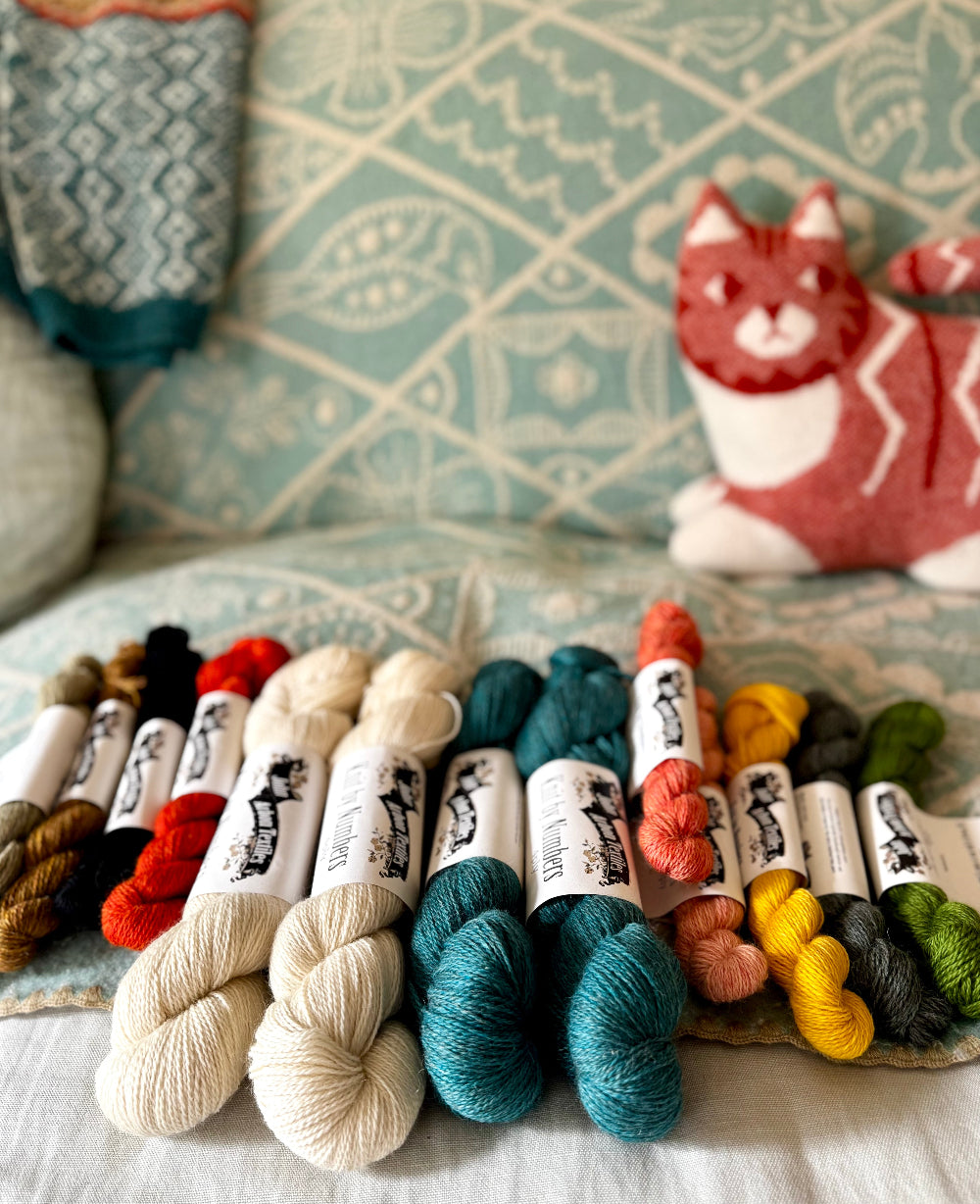Assorted skeins of yarn on a textured surface with a cat-themed pillow in the background.