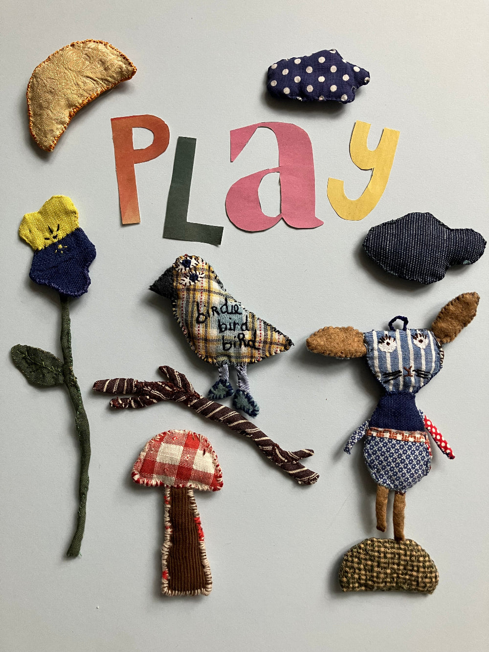 A collection of handmade, patchwork-style fabric toys and decorative cutouts arranged on a light gray background. The set includes a rabbit, a mushroom, a flower, a crescent moon, a ribbon, and the word 'play' spelled out in letters, all crafted from various patterned fabrics.