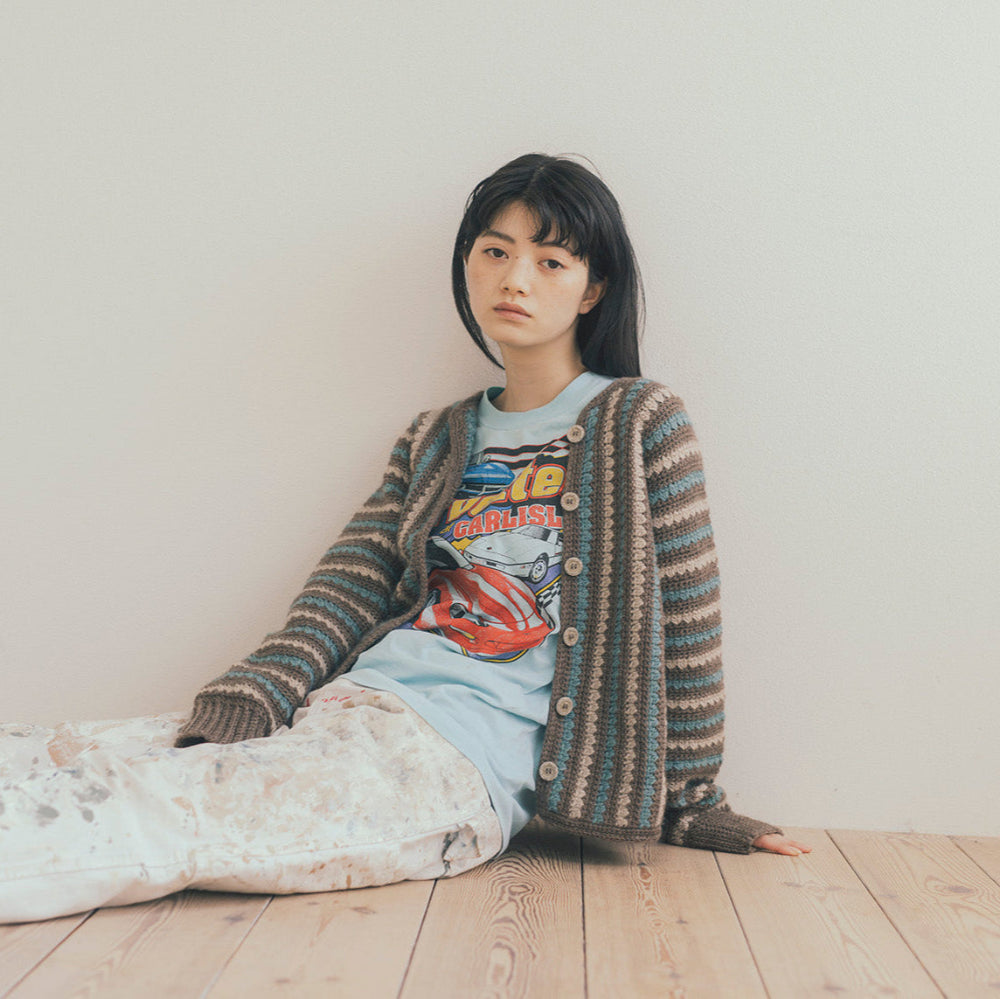 Person sitting on a bed wearing a patterned cardigan and graphic t-shirt against a plain wall.
