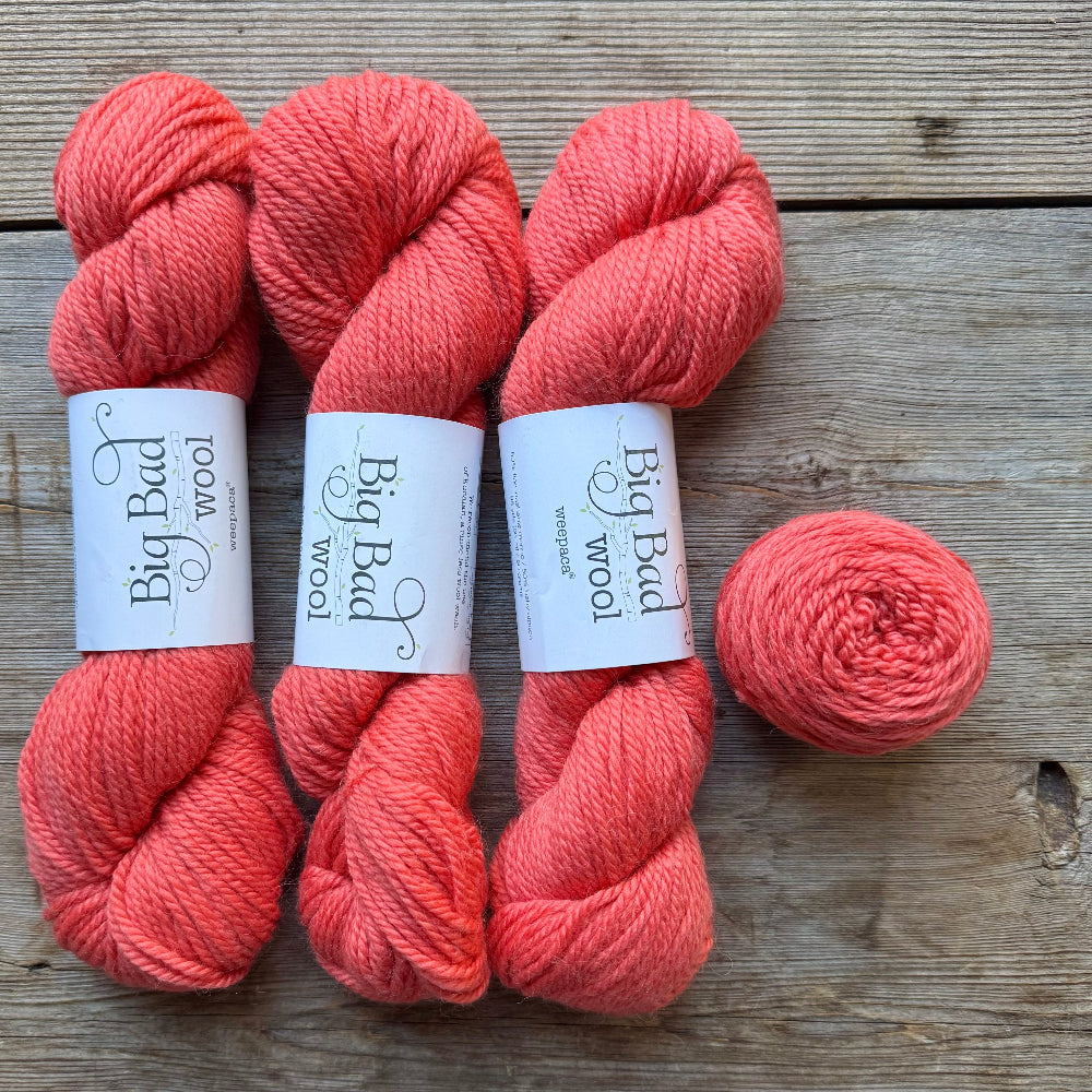Three skeins of coral pink yarn and one matching ball of yarn laid on a rustic wooden surface. Each skein has a white paper label reading Big Bad Wool.