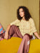 Woman sitting on a yellow couch wearing a beige crochet top and brown pants.