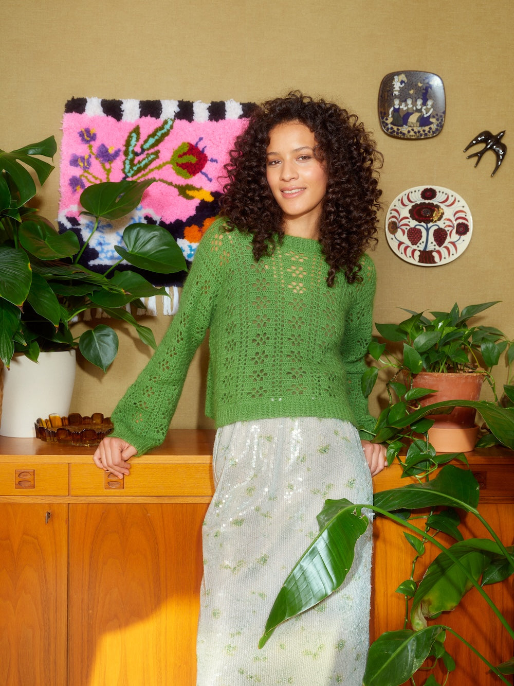 Woman in a green sweater standing in a room with plants and decorative items on the wall.