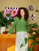 Woman in a green sweater standing in a room with plants and decorative items on the wall.