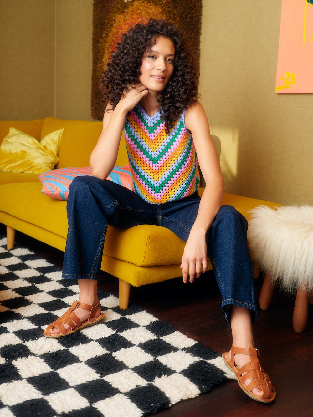 Woman wearing a colorful crocheted vest sitting on a yellow couch in a room with a checkered rug.