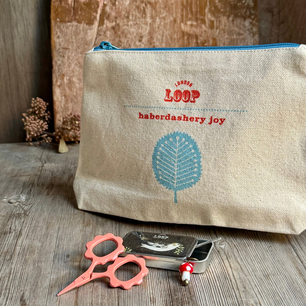 A natural linen cosmetic pouch with a blue zipper, featuring red embroidered text reading 'LOOP haberdashery joy' and a blue leaf design. The pouch is styled on a rustic wooden surface with pink-handled scissors and a small tin of pearl beads in front of it.