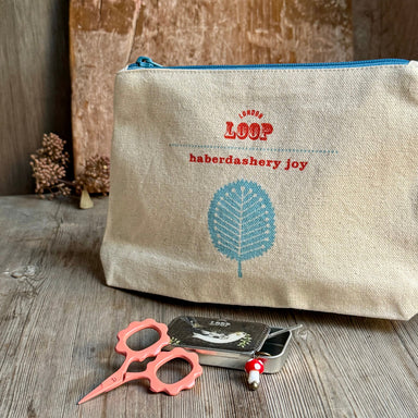 A natural linen cosmetic pouch with a blue zipper, featuring red embroidered text reading 'LOOP haberdashery joy' and a blue leaf design. The pouch is styled on a rustic wooden surface with pink-handled scissors and a small tin of pearl beads in front of it.