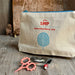 A natural linen cosmetic pouch with a blue zipper, featuring red embroidered text reading 'LOOP haberdashery joy' and a blue leaf design. The pouch is styled on a rustic wooden surface with pink-handled scissors and a small tin of pearl beads in front of it.