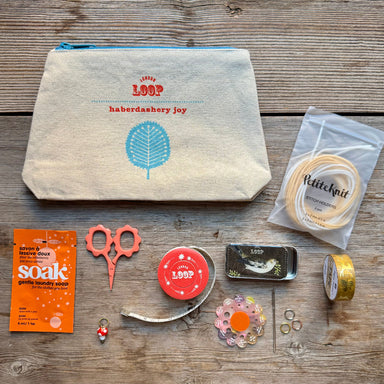 A flat-lay of a sewing and knitting kit on a rustic wooden background, featuring a canvas pouch, knitting needles, yarn, scissors, soap, thread, ribbon, and other accessories. The brand 'London Loop' is visible on the pouch, soap packet, and tin.