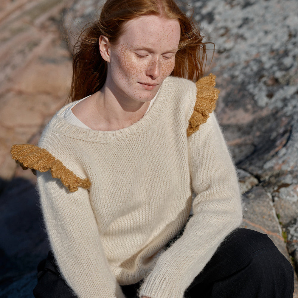 Woman wearing a cream sweater with ruffled shoulders sitting on rocks.
