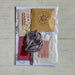 A packaged fabric and textile kit containing a ball of dusty purple ribbon, a pink lace trim, a beige fabric swatch, a red fabric swatch, a white fabric swatch, and a kraft paper envelope with a printed logo, all laid out on a light wood background.