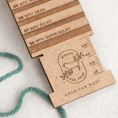 A close-up of a wooden measuring tool for yarn wraps, showing engraved size labels from 06 to 14 and a sheep logo with the text 'WRAPS PER INCH' and 'AMERICAN MADE.' A mint green yarn tail is draped in the lower left on a light fabric background.