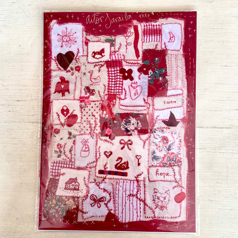 A square fabric art patchwork sampler card or kit in clear plastic packaging, featuring a red border and various stitched designs in red, pink, and white on a light wood background.