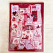 A square fabric art patchwork sampler card or kit in clear plastic packaging, featuring a red border and various stitched designs in red, pink, and white on a light wood background.