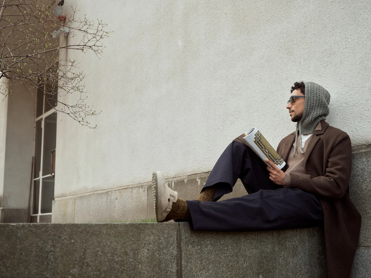 Man sitting on a ledge reading a book against a plain wall.