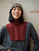 Person wearing a gray sweater with a red knitted turtleneck and a knit beanie against a textured wall.