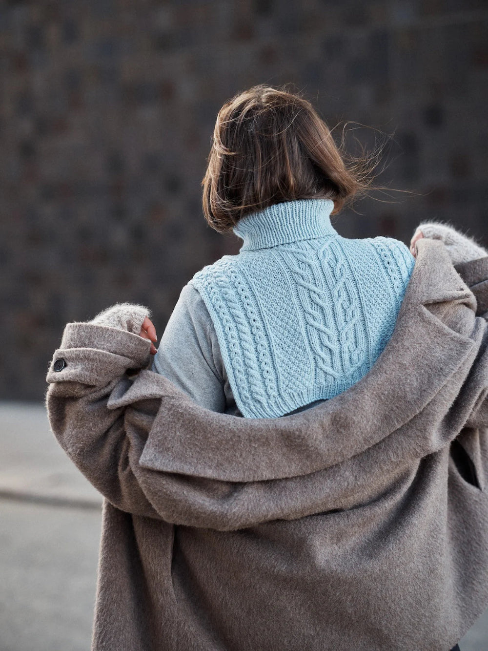 Person wearing a light blue knitted sweater with a brown coat over it, standing against a dark background.