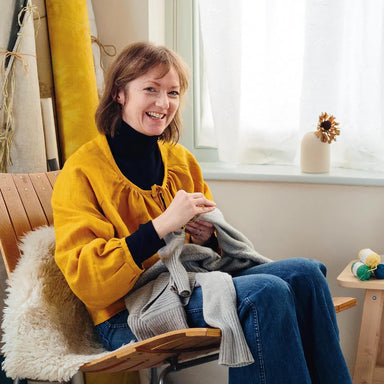 Woman sitting on a wooden chair wearing a yellow sweater and blue jeans, holding a gray sweater.
