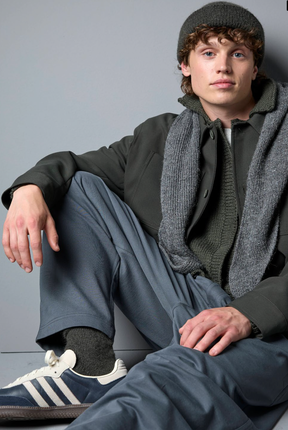 Person wearing a dark coat, gray scarf, and Adidas sneakers against a gray background